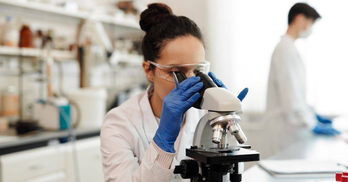 A female scientist in protective gear examines samples through a microscope in a laboratory setting.