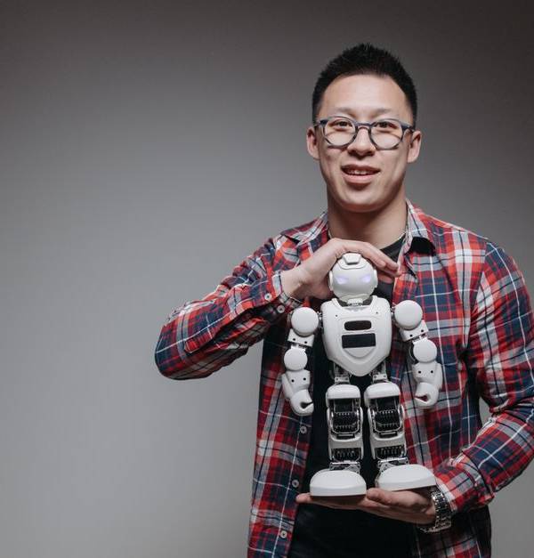 Asian man with eyeglasses holding a toy robot in a studio with a gray background.