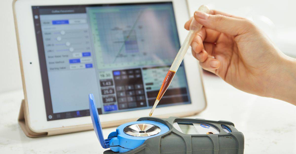 Hand using pipette with refractometer in front of tablet displaying measurement interface.